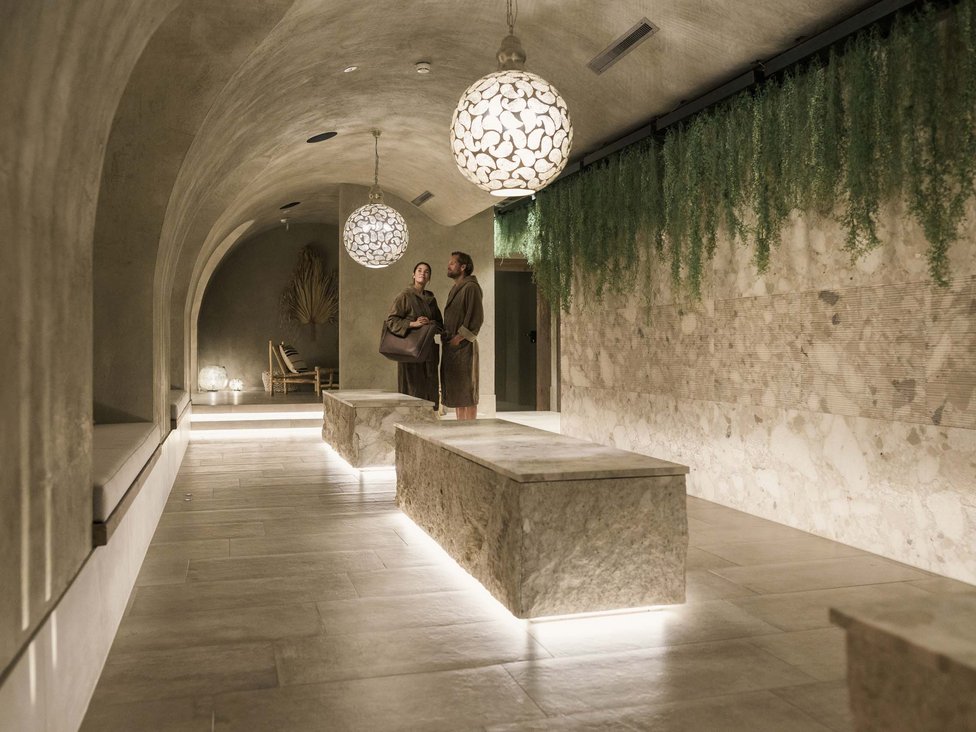 Großzügig und exklusiv: Ihr Wellnessurlaub in Tirol Zwei Personen in Bademänteln in einem modern beleuchteten Spa-Flur mit Steinbänken