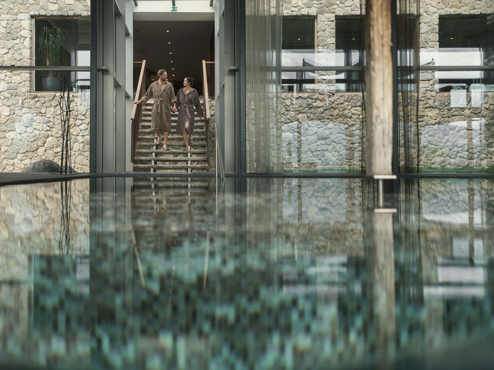 Großzügig und exklusiv: Ihr Wellnessurlaub in Tirol Paar in Bademänteln geht eine Treppe neben einem Innenpool in einem Spa