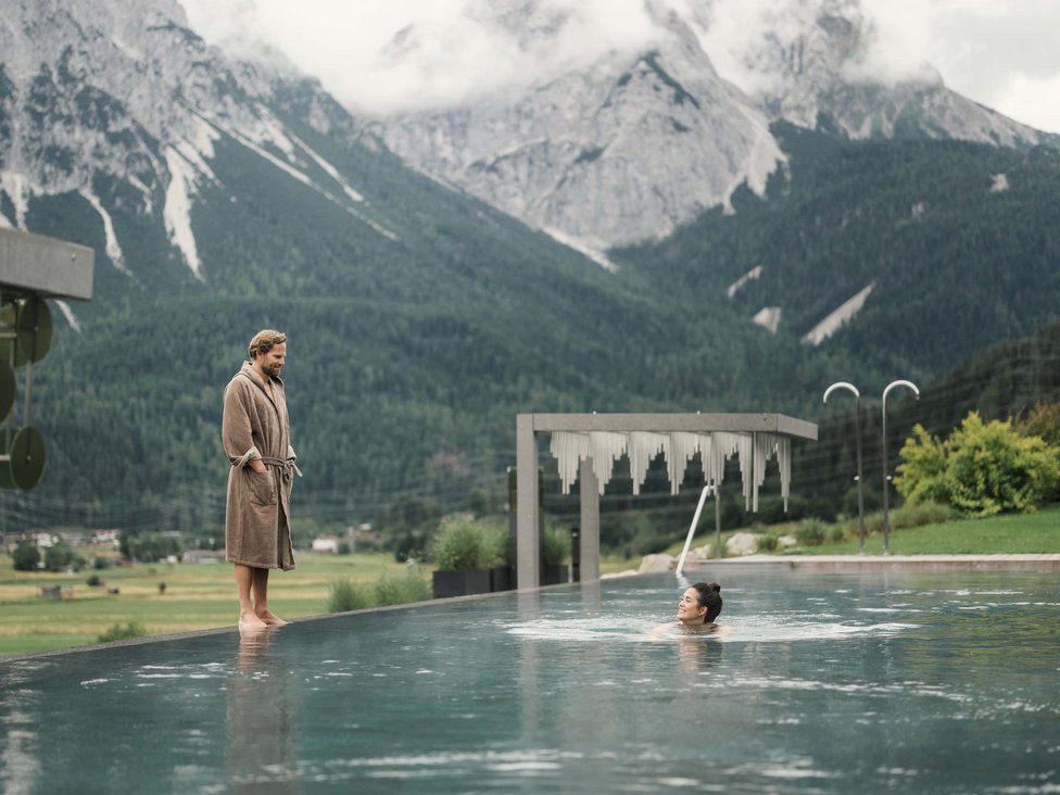 Großzügig und exklusiv: Ihr Wellnessurlaub in Tirol Mann im Bademantel steht am Pool, Frau schwimmt, Berge im Hintergrund