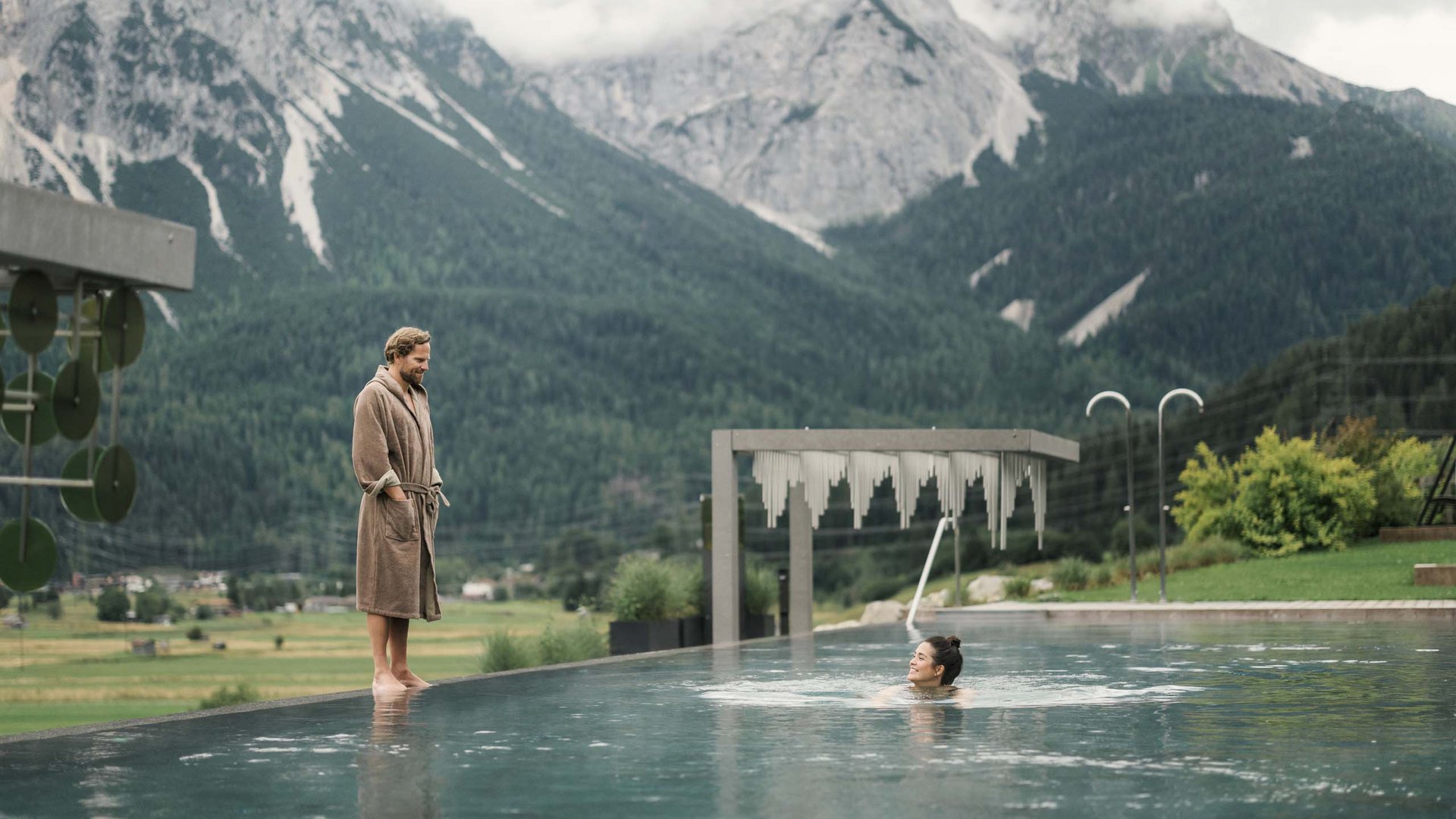 MOHR life resort Bildergalerie Mann im Bademantel steht am Pool, Frau schwimmt, Berge im Hintergrund