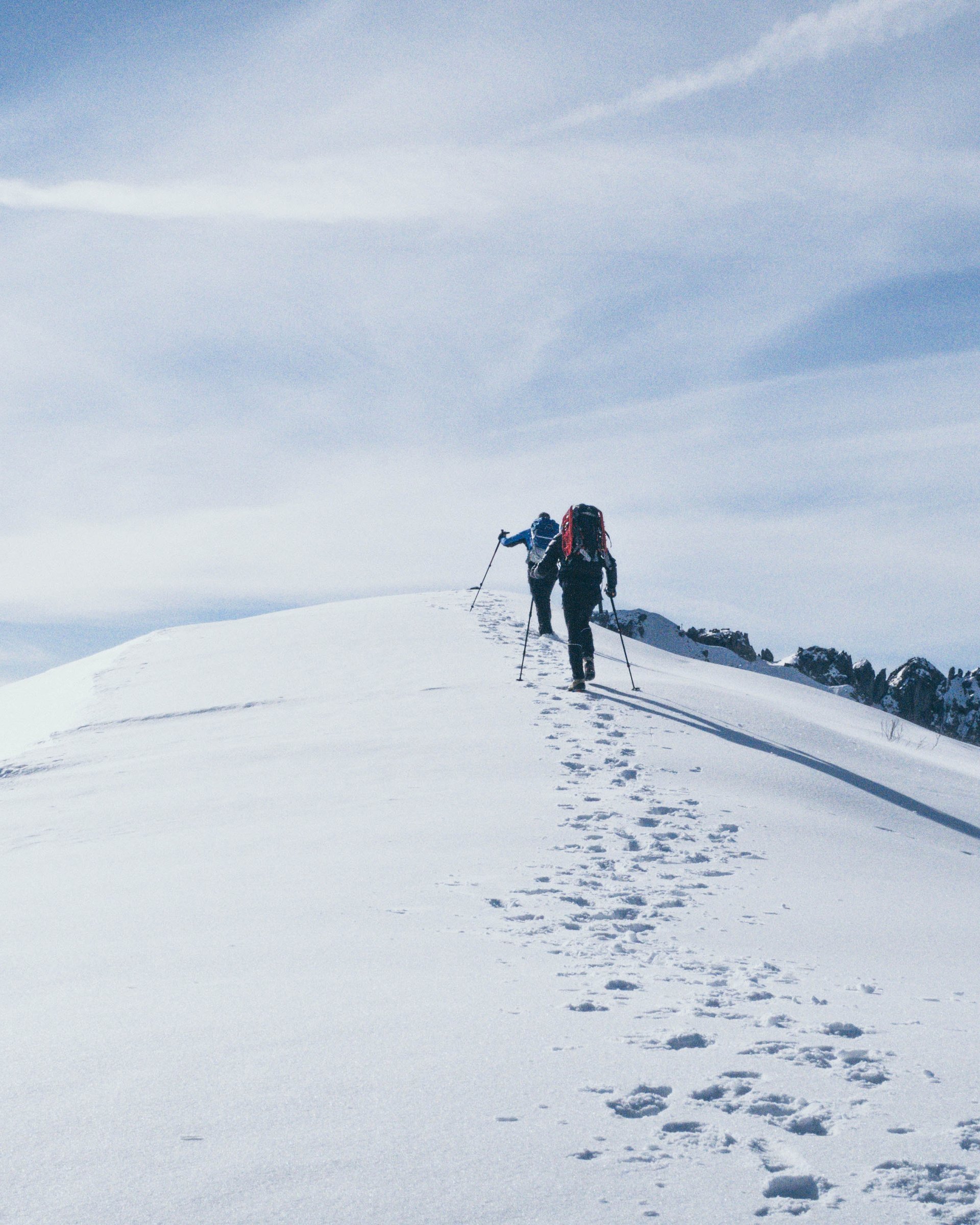 Rodeln in Lermoos – macht Spaß! Zwei Wanderer steigen mit Stöcken auf eine schneebedeckte Bergspitze bei blauem Himmel