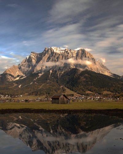 Ihr Favorit? MOHR life resort – das Hotel in Lermoos Berg mit Wolken und Spiegelung im Wasser vor einer Hütte und Dorf