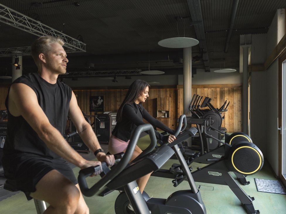 Wellness und Luxus: Ihr Hotel in Tirol Mann und Frau trainieren auf Fahrradergometern im Fitnessstudio mit Holzverkleidung