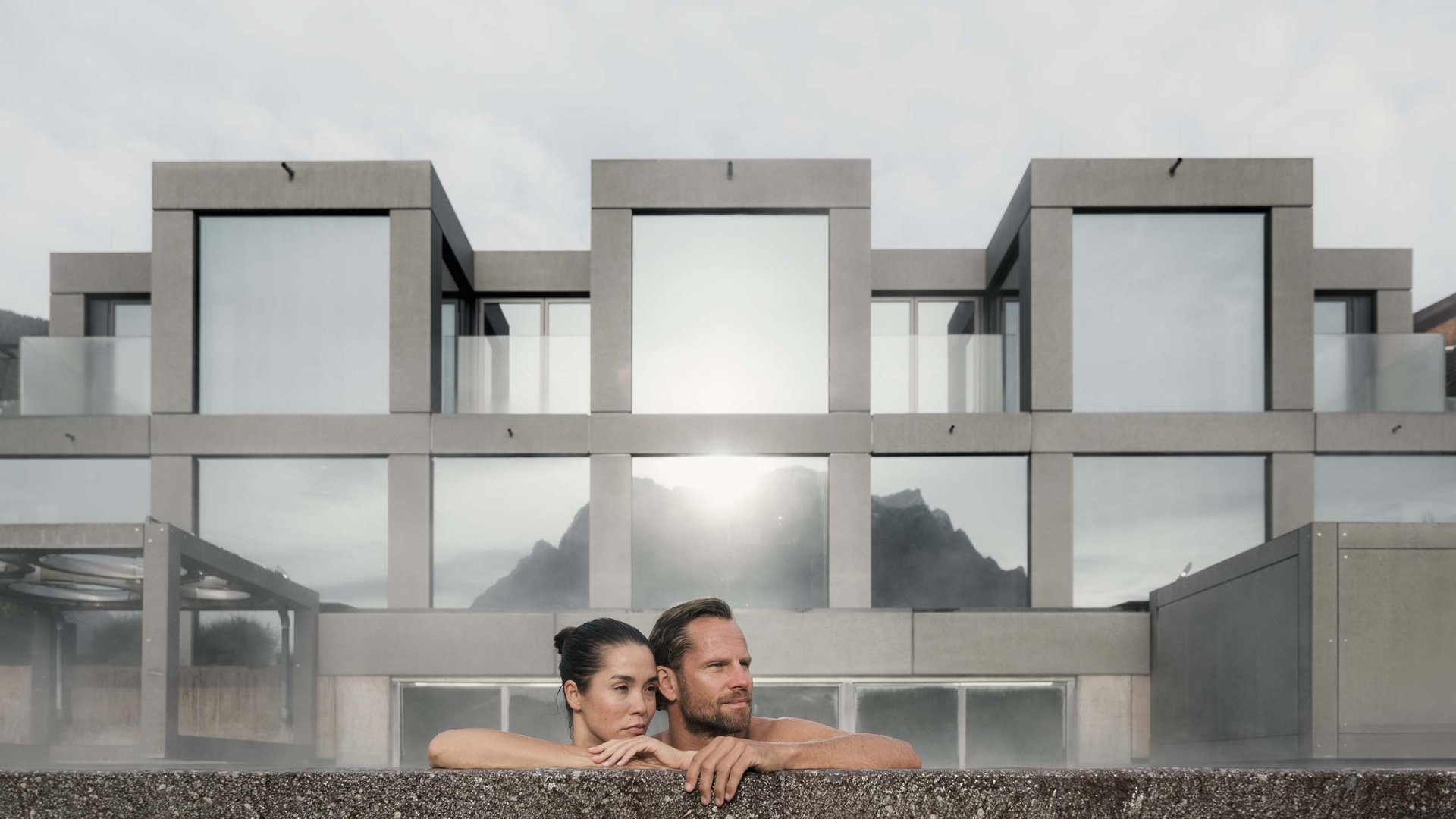 MOHR life resort Bildergalerie Paar entspannt in beheiztem Außenpool vor modernem Gebäude mit Bergblick