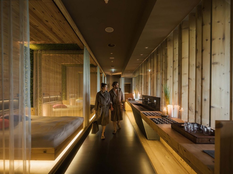 Großzügig und exklusiv: Ihr Wellnessurlaub in Tirol Ein Paar in Bademänteln geht durch einen warm beleuchteten Spa-Flur aus Holz.