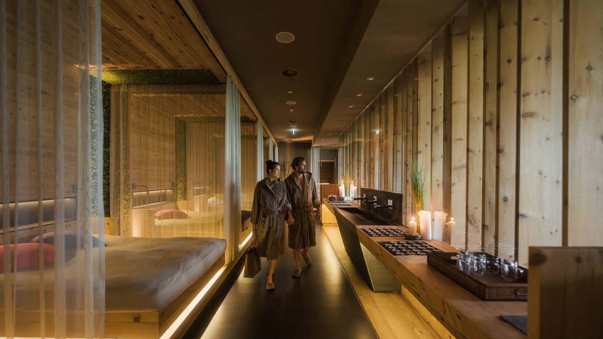 MOHR life resort Bildergalerie Ein Paar in Bademänteln geht durch einen warm beleuchteten Spa-Flur aus Holz.