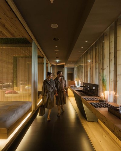MOHR life resort – wunderbares Wellnesshotel in Lermoos Ein Paar in Bademänteln geht durch einen warm beleuchteten Spa-Flur aus Holz.