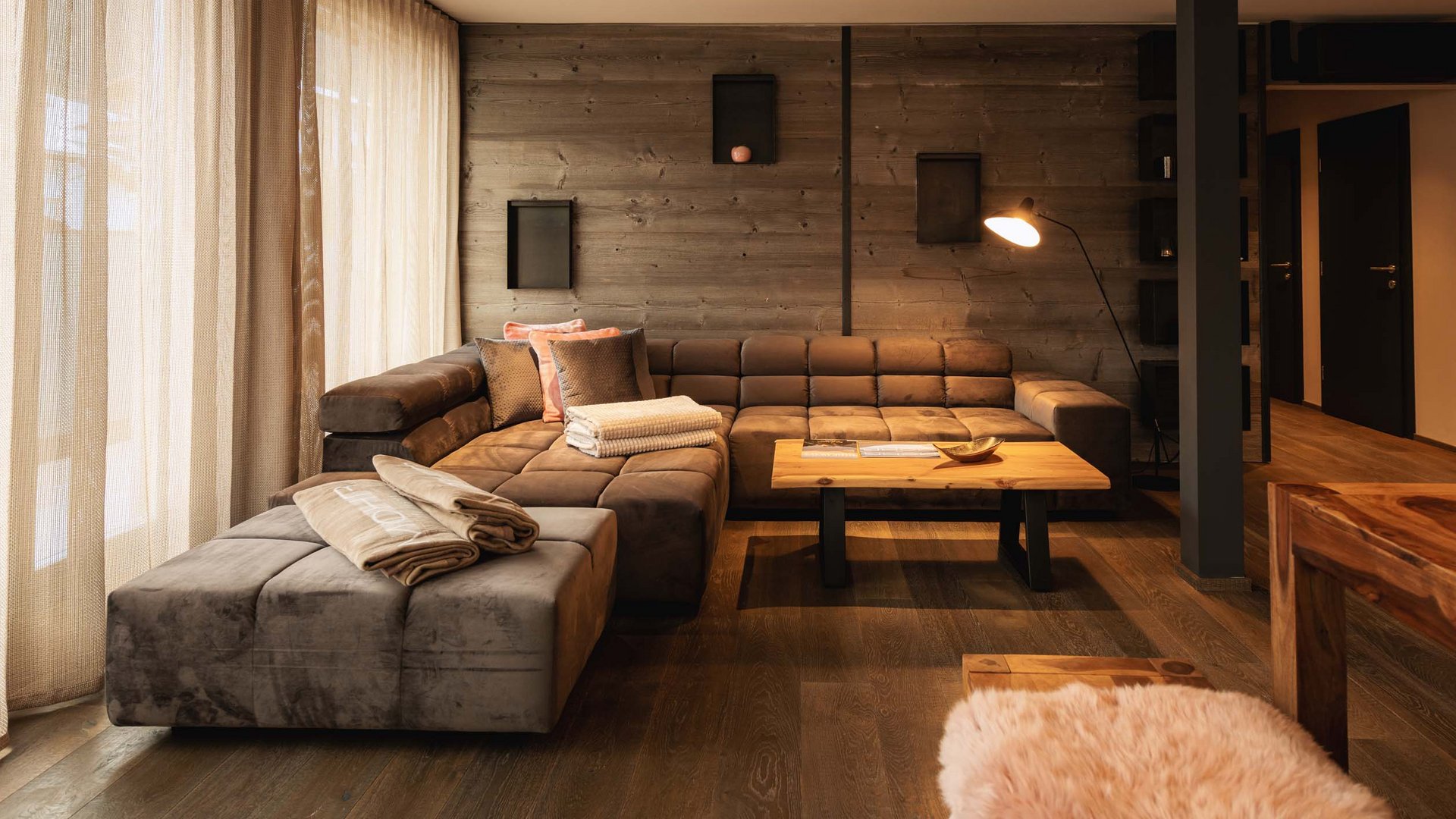 Wellness und Luxus: Ihr Hotel in Tirol Gemütliches Wohnzimmer mit braunem Ecksofa, Holztisch und weichen Vorhängen