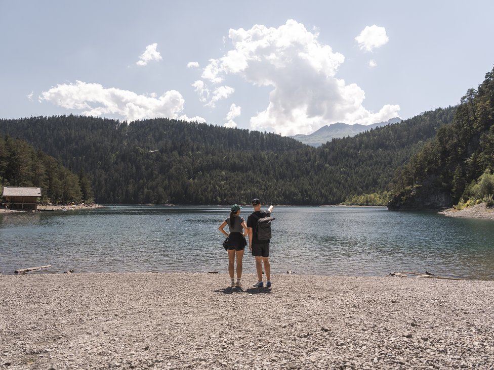Wellness und Luxus: Ihr Hotel in Tirol Zwei Personen stehen am Kiesstrand und schauen auf einen See mit bewaldeten Bergen