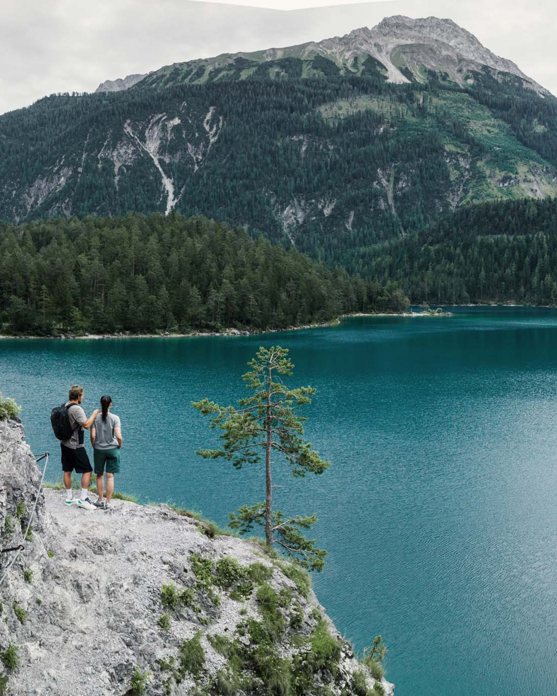 Auf Tour mit MOHR Zwei Personen wandern an einem See mit Bergen und Wald im Hintergrund