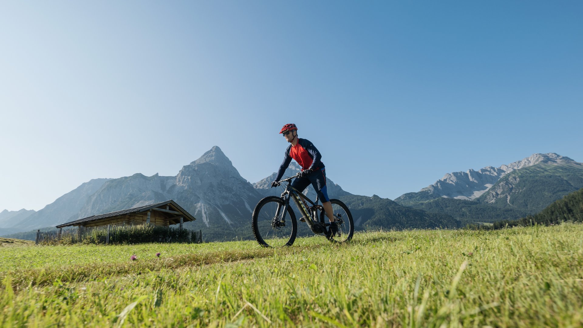 Auf Tour mit MOHR Mann fährt Mountainbike auf einer Wiese mit Bergkulisse bei klarem Himmel