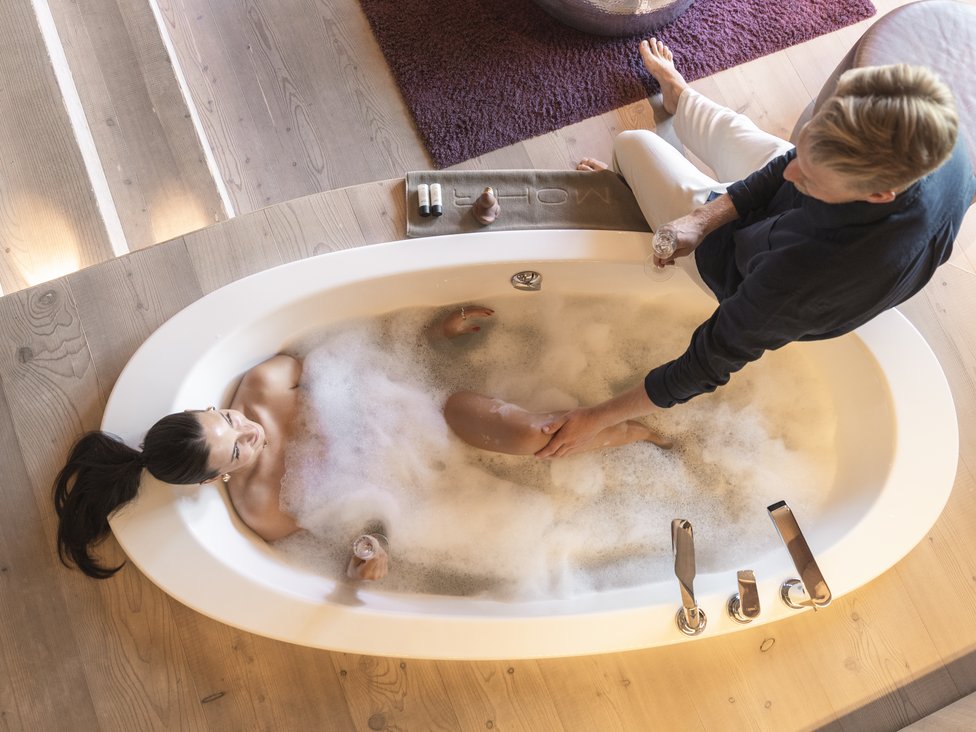 Wellness und Luxus: Ihr Hotel in Tirol Frau entspannt in Schaumbad, Mann sitzt neben Badewanne und hält ihr Bein