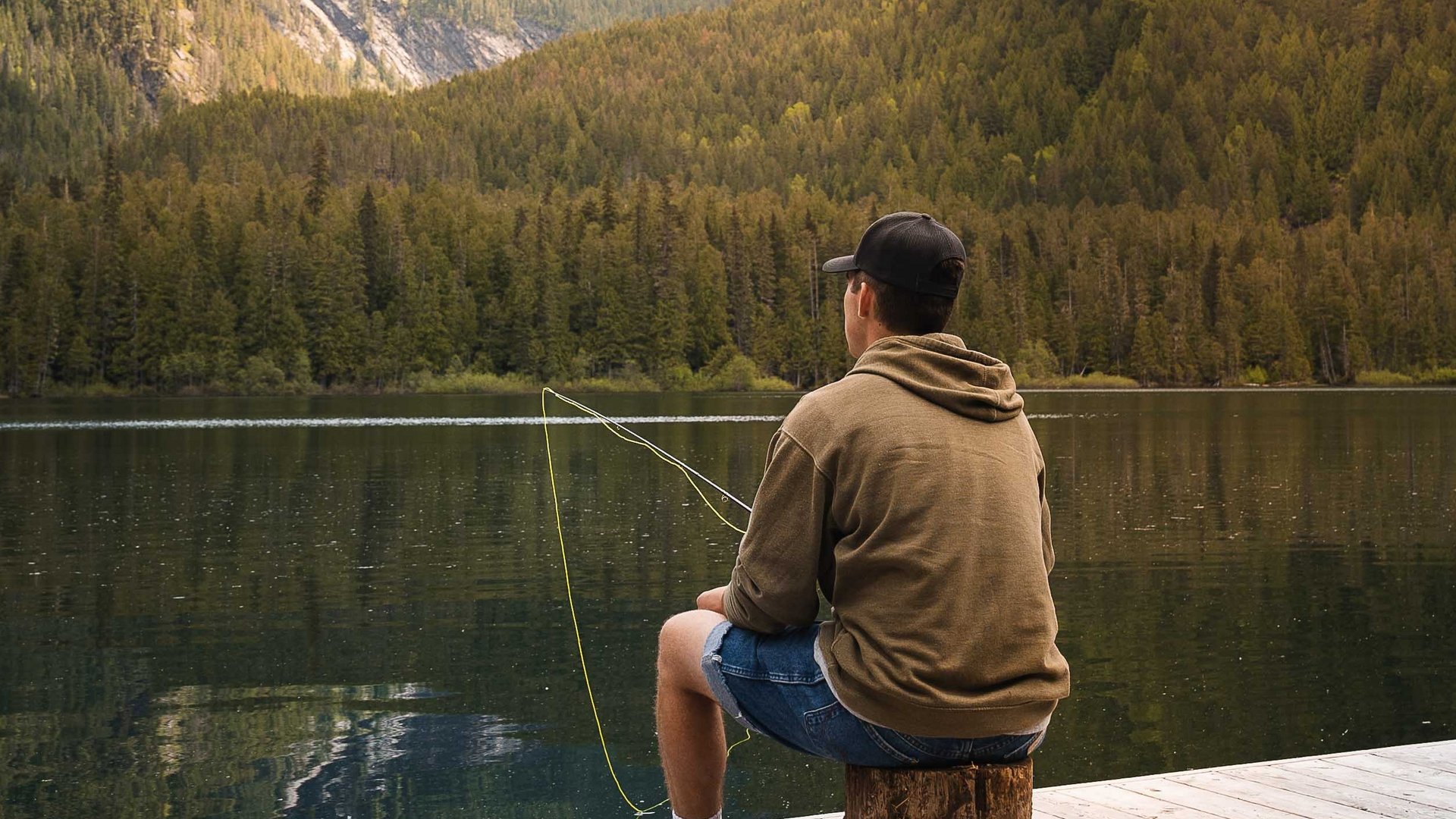 Fischerfun in Lermoos Mann sitzt am Steg und angelt in einem Bergsee vor bewaldeten Bergen