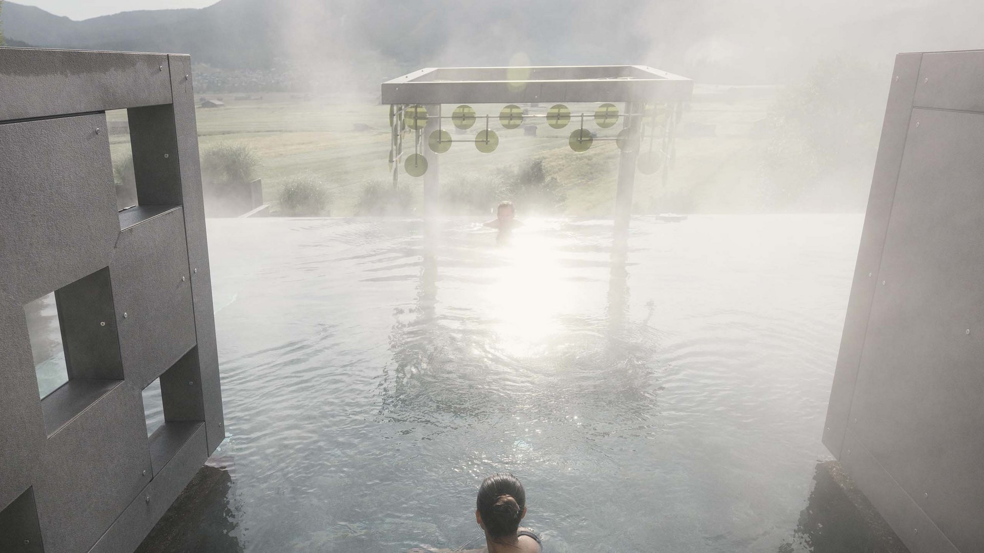 MOHR life resort Bildergalerie Person betritt dampfendes Außenbecken mit Bergpanorama im Sonnenlicht