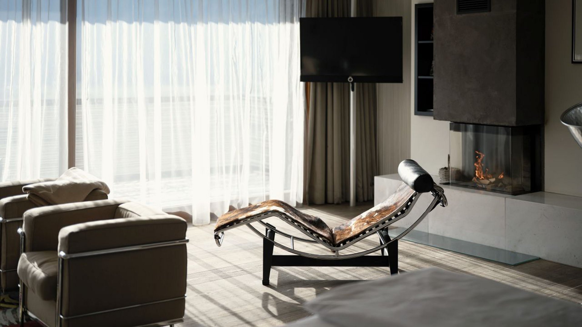 MOHR life resort Bildergalerie Moderner Wohnbereich mit Ledersesseln, Liege und Kamin vor großen Fenstern