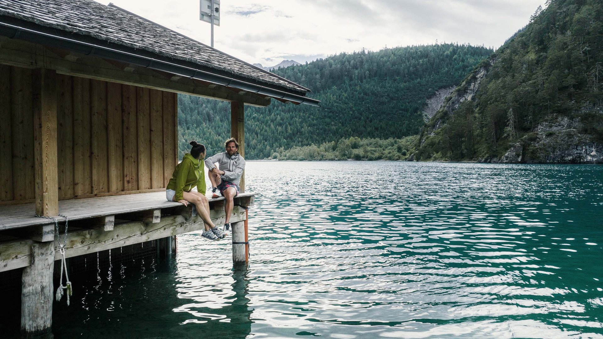 Bereit für MOHR? Das Sporthotel in Lermoos Zwei Personen sitzen auf einem Steg neben einem See mit bewaldeten Bergen